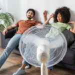 couple feeling warm and exhausted in front of an electric fan due to AC not keeping up