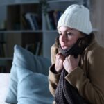 Woman covered with winter clothes feeling cold while sitting on the couch due to furnace failure