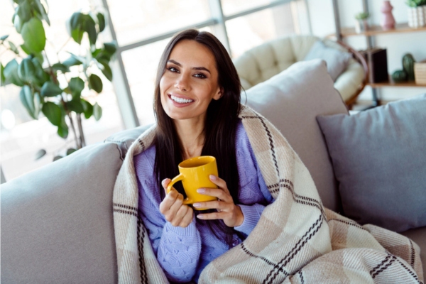 woman smiling while drinking coffee on the couch wrapped in blanket depicting Weather-Ready Protection All Winter